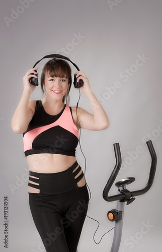 Woman holding headphones with an exercise bicycle against a grey background