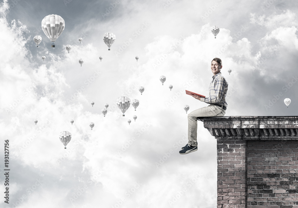 Man on roof edge reading book and aerostats flying in sky Man on roof edge reading book and aerostats flying in sky