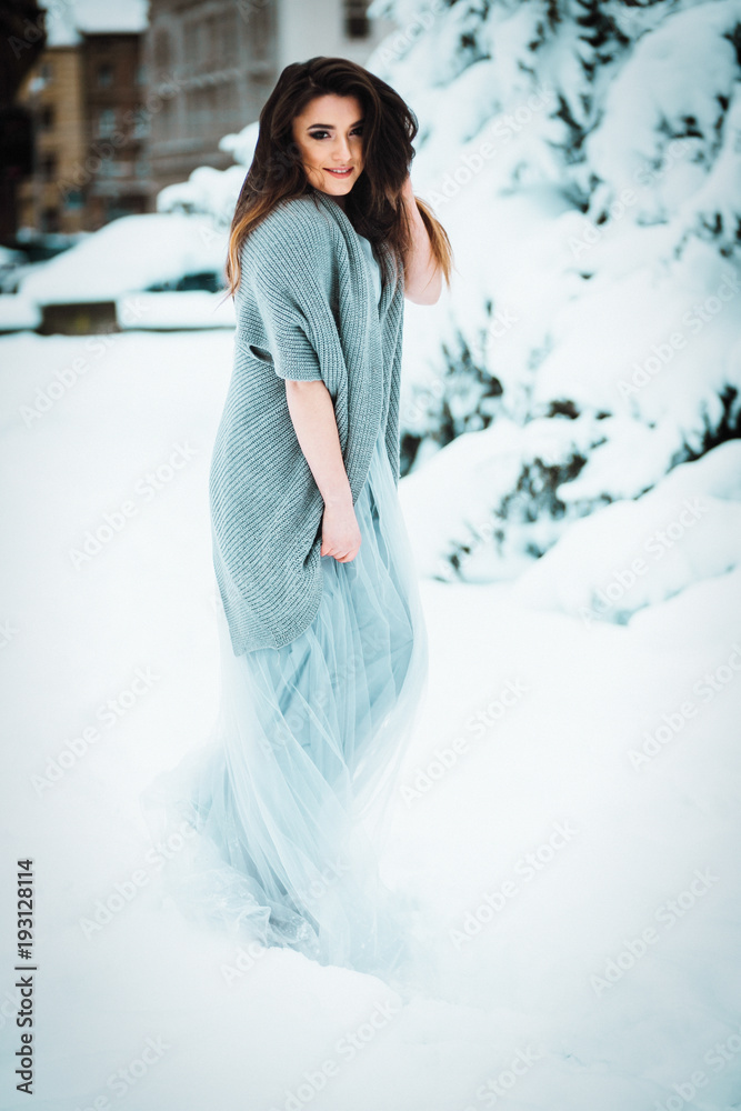 Stunning woman in a sweater and light dress stands in the garden among bushes covered with snow Stunning woman in a sweater and light dress stands in the garden among bushes covered with snow