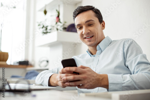 In high spirits. Attractive alert neat dark-haired man smiling and typing on his phone while sitting at the table