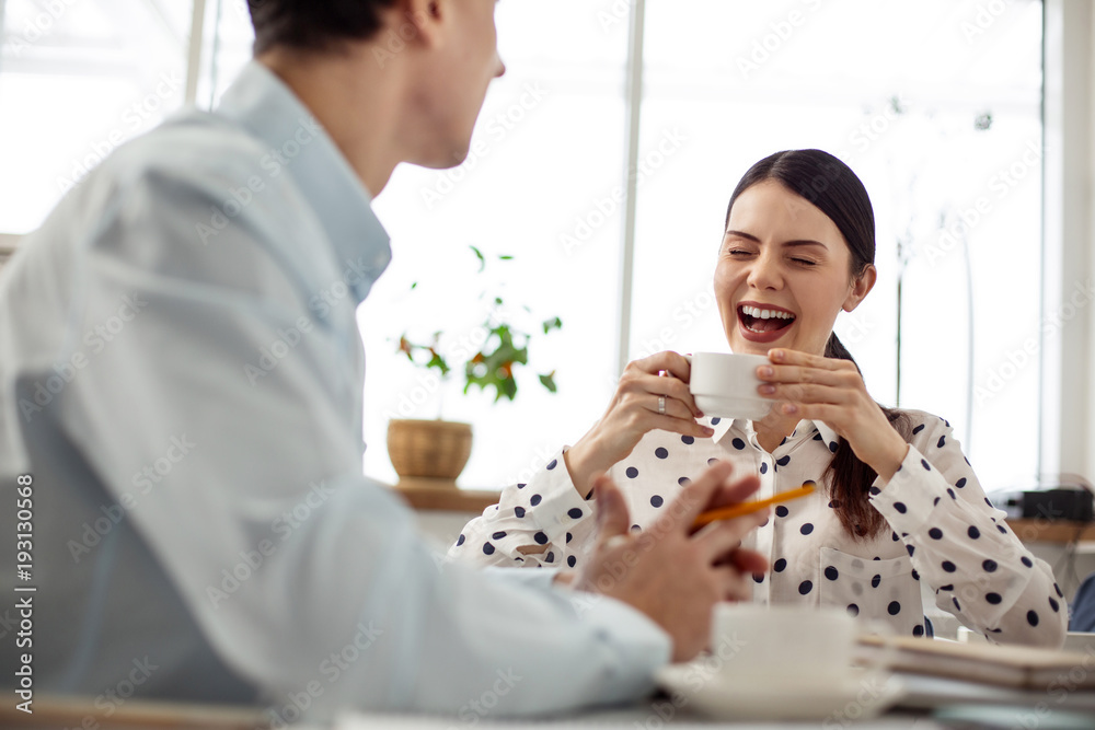 Laughing. Pretty overjoyed dark-haired young woman laughing and having a cup of coffee while talking to her colleague Laughing. Pretty overjoyed dark-haired young woman laughing and having a cup of coffee while talking to her colleague