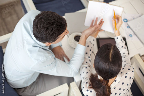 My colleague. Neat determined dark-haired man talking with his female colleague sitting next to him and she holding a notebook