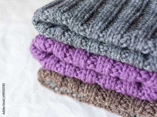 A pile of handmade knitted hats of gray, lilac and brown colors on white bokeh background close up