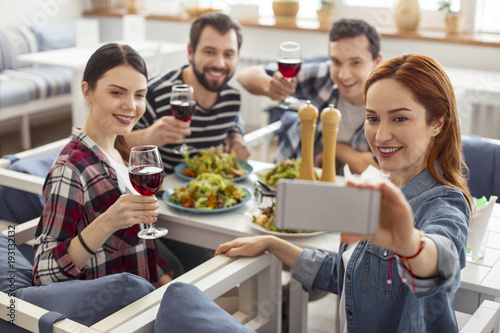 Selfie time. Good-looking happy old best friends smiling and sitting together at the table and drinking wine and one woman taking a selfie