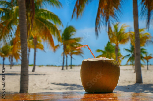 Coconut with a straw on the table on a background of palm trees and tropical sand beach
