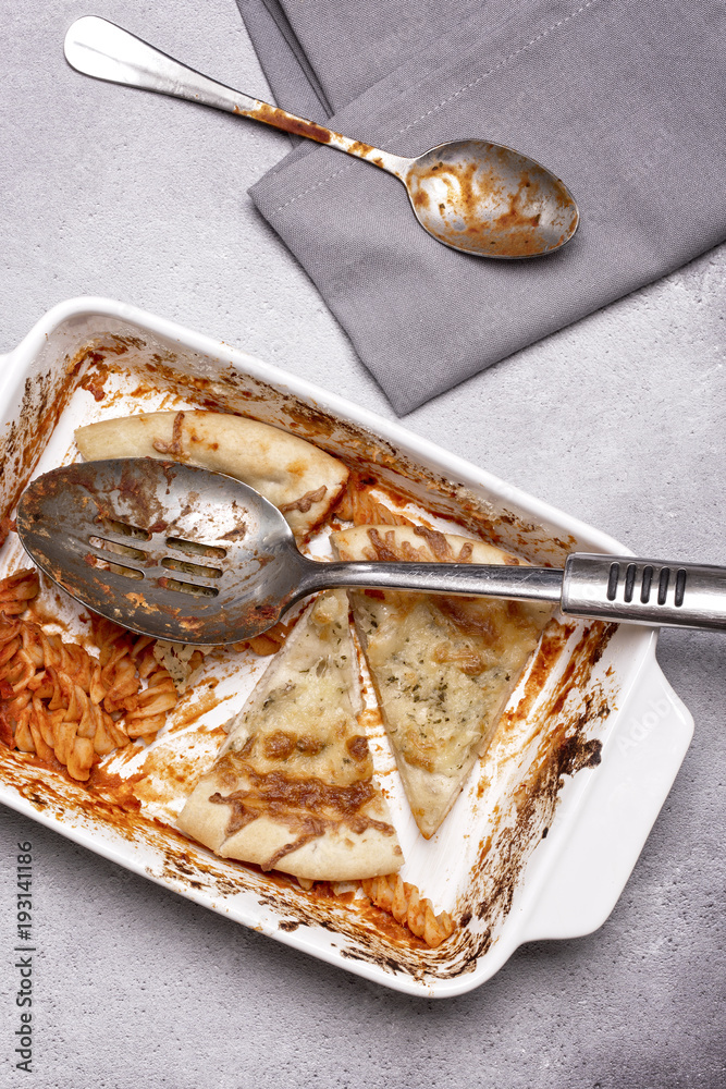 Fototapeta premium Dish of leftover pasta bake and cheesy garlic bread with a spoon and napkin on a grey rustic background
