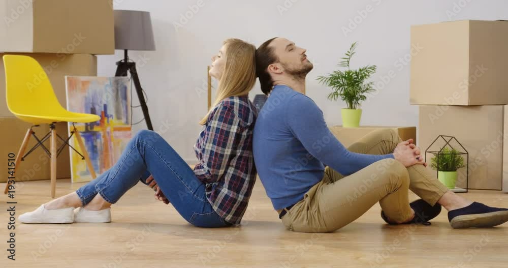 Young attractive couple sitting back to back on the floor of the cozy light room and resting among packed boxes after moving in new home. Inside Young attractive couple sitting back to back on the floor of the cozy light room and resting among packed boxes after moving in new home. Inside