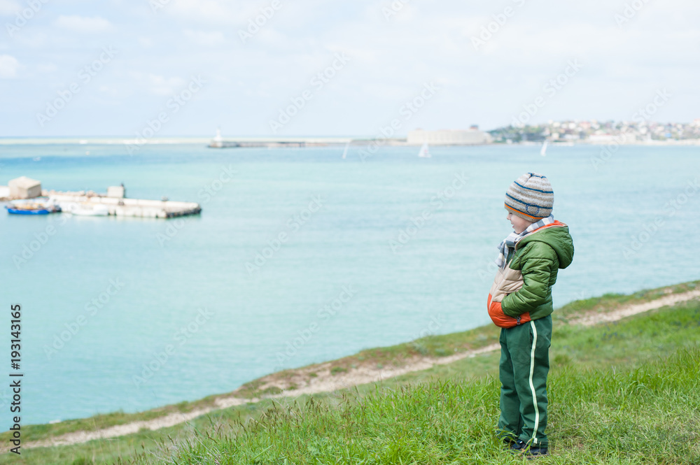 lonely little boy in jacket and hat stands on slope against background of sea in spring