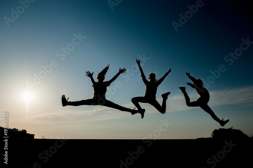 three silhouettes of jumping teen girls on blue sky background at sunset