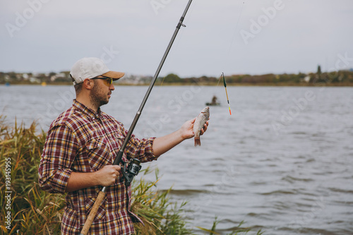 Side view Young unshaven man in checkered shirt, cap, sunglasses pulls out fishing pole and holds caught fish on shore of lake near shrubs and reeds. Lifestyle, recreation, fisherman leisure concept