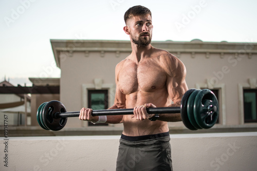 Muscular man with perfect abs, shoulders, biceps, triceps and chest workout with barbell at outdoor gym.