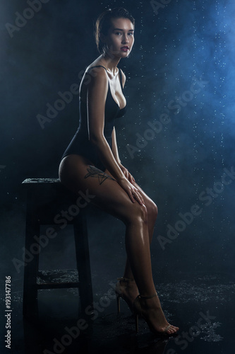Beautiful wet big breasted girl wearing black swimsuit sits on a chair on a dark background. Falling rain drops and artistic scenic smoke