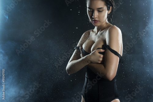 Beautiful wet big breasted girl wearing black swimsuit on a dark background. Falling rain drops and artistic scenic smoke