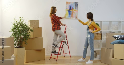 Handsome young boyfriend standing on the ladder and hanging a painting on the wall while his charming girlfriend rulling him among unpacked stuff during moving in together. Indoor photo