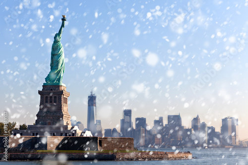 The Statue of Liberty free of tourists and New York City Downtown on sunny early morning during snowfall.