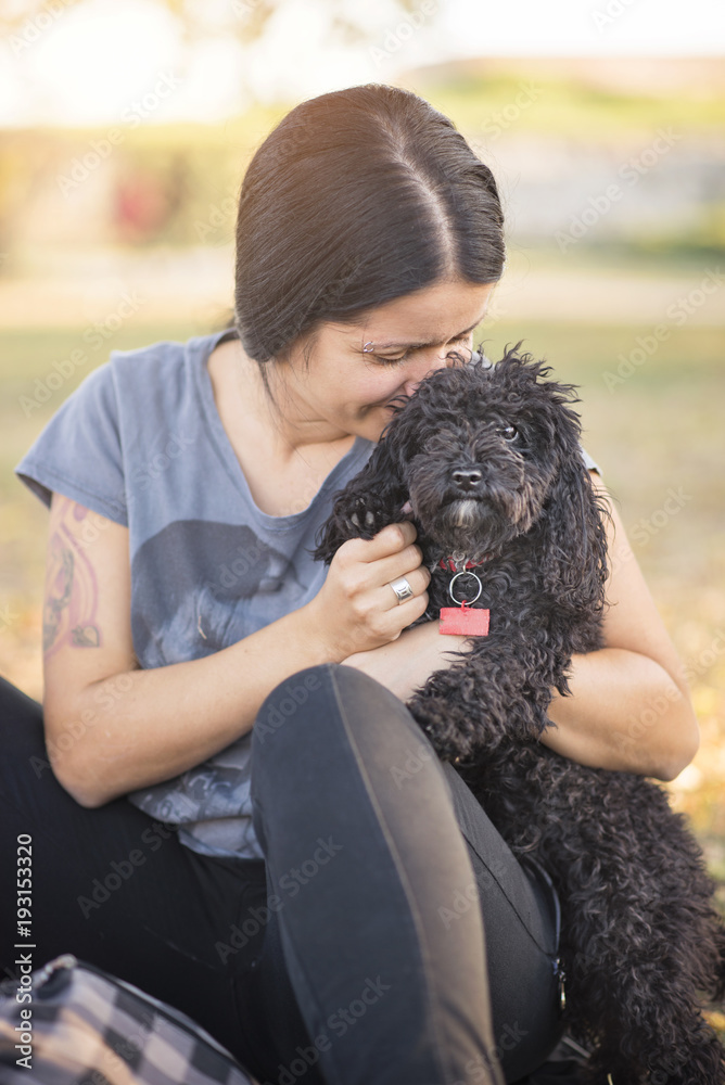 Young woman cuddling with her cute little miniature black poodle, sitting together in a park, enjoying the sunny and warm weather