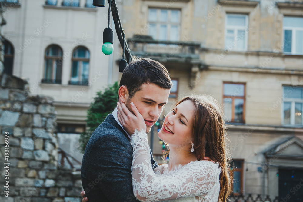 Wedding couple hugs in the old city. Rustic bride with hair down holds face of a groom in grey suit and bow tie. Love in ancient medieval town. White lace wedding dress. Vintage architecture details.