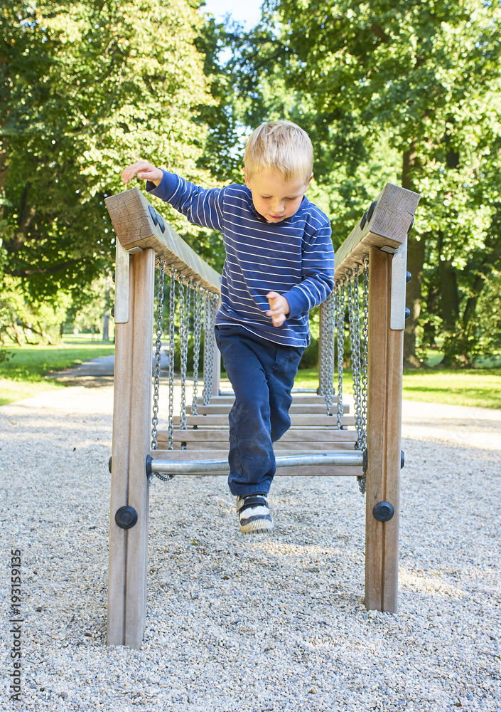 The boy is on the rope bridge in the Playground. The child carefully steps  over the gaps in the hanging bridge. The boy on the Playground in summer  Photos