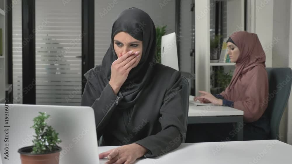 Girl in black hijab sits in the office and coughs, a disease, a cold, an illness is a concept. Girl in pink hijab in the background. 60 fps Girl in black hijab sits in the office and coughs, a disease, a cold, an illness is a concept. Girl in pink hijab in the background. 60 fps