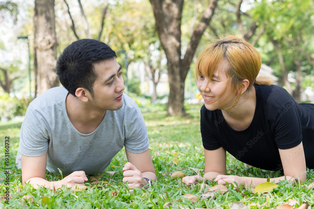 Asian sweet couple exercising together with smile on their faces on soft grass encircle with nature and warm light from the sun in the afternoon. Couple smile happily to each other. Outdoor exercise. Asian sweet couple exercising together with smile on their faces on soft grass encircle with nature and warm light from the sun in the afternoon. Couple smile happily to each other. Outdoor exercise.