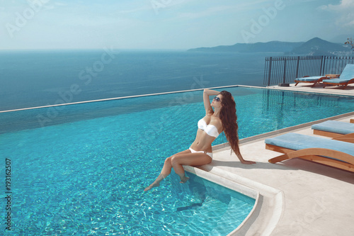 Sexy model in white bikini sunbathing by infinity swimming pool. Luxury travel vacation. Brunette Woman sunbathing at luxurious resort. Beautiful destinations concept. 