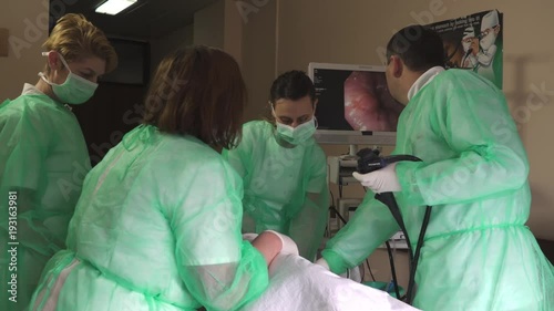 Medical team examining colon of patient who lying on bed, gastroenterologist with joystick control micro camera for colonoscopy and looking on computer screen, nurses in uniforms assisting doctor.