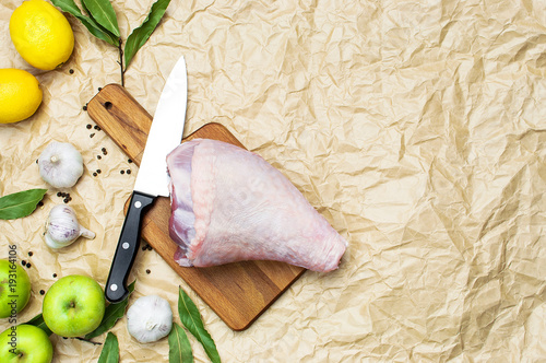 Raw turkey thigh garlic apples lemon quail eggs spices on a wooden board. Top view of a turkey, knife on a beige paper background. Culinary background, ingredients for cooking. photo