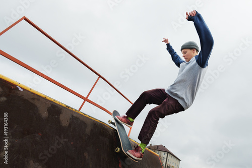 A teenager skateboarder in a hat does a Rocks trick on a ramp in a skate park against a cloudy sky and sleeping area. The concept of urban style in sport