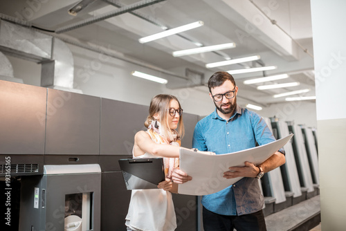 Young woman designer and print operator working together with paper print standing at the print manufacturing