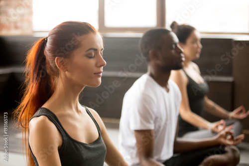 Group of young sporty afro american and caucasian people practicing yoga lesson  sitting in Sukhasana exercise  Easy Seat pose  working out  students training in sport club  studio close up