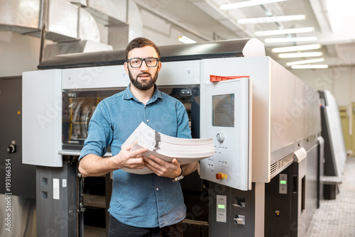 Portrait of a handsome typographer standing with stack of paper at the printing manufacturing with offset machine on the background