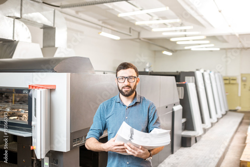 Portrait of a handsome typographer standing with stack of paper at the printing manufacturing with offset machine on the background
