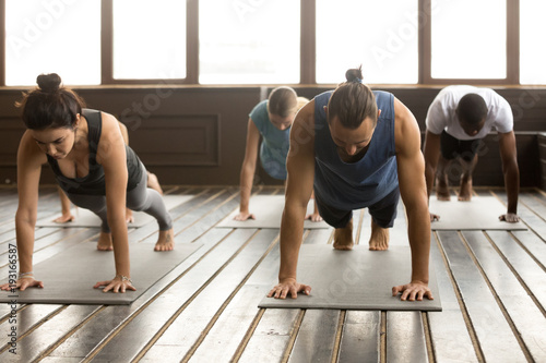 Group of young yogi people practicing yoga lesson standing in Plank pose, doing Push ups or press ups exercise, working out, indoor full length, studio floor