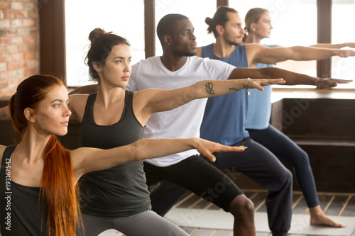 Group of young sporty afro american and caucasian people practicing yoga lesson, standing in Warrior two exercise, Virabhadrasana II pose, working out, indoor close up, studio background