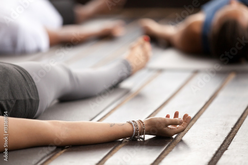 Foto Group of young sporty people practicing yoga, lying in Corpse pose, Savasana exercise, working out, resting after practice, female hand with wrist bracelets close up, studio
