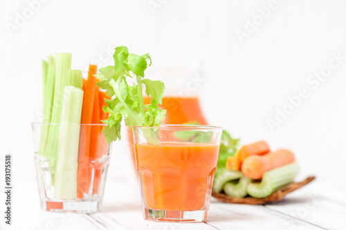 Carrot juice in beautiful glasses, cut carrot and green celery on wood bark bowl on white wooden background. Fresh vegetable drink. Close up photography. Selective focus. Horizontal banner