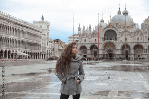 Beautiful tourist girl with long wavy hair in San Marco square in front of St. Mark Basilica, Venice, Italy. Happy smiling brunette wearing in gray coat enjoying holiday.