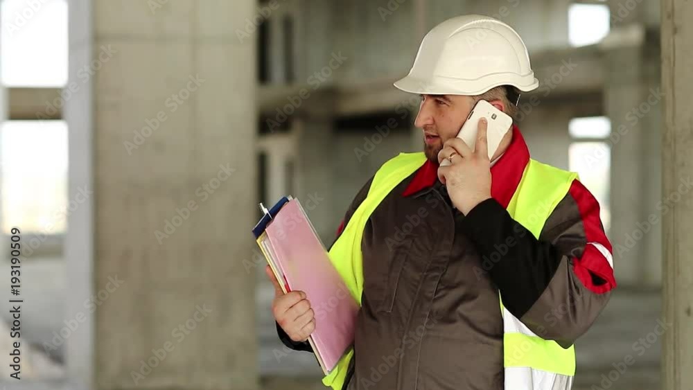 Builder with smartphone at construction site. Foreman with work documentation in yellow hard hat on construction site talks on smartphone. Builder speaks on cell phone on project site Builder with smartphone at construction site. Foreman with work documentation in yellow hard hat on construction site talks on smartphone. Builder speaks on cell phone on project site