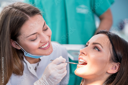 people, medicine, stomatology and health care concept - happy female dentist with mirror checking patient girl teeth up at dental clinic office while male assistant is waiting behind.