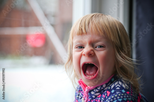 Close-up Portrait of Cute Blonde 18 Month Old Toddler Girl with Big Blue Eyes, Angry, Crying, Screaming Face Expression, Stand by the Window, Snowy Background 