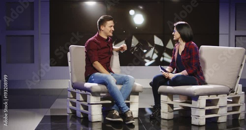 Young confident and relaxed man and woman taking participation in television show sitting in modern illuminated studio and having talk.