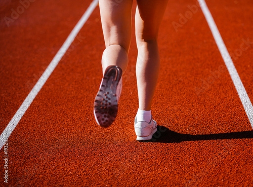 Young girl legs in white sneakers back side on a red race track of a stadium with white lane lines on background. © andrew_shots