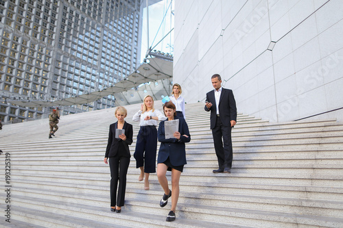 Employees of large  corporation walking in   and speaking by smartphone. Concept of successful business partners outside. Young prosperous people dressed in black suits look happy.