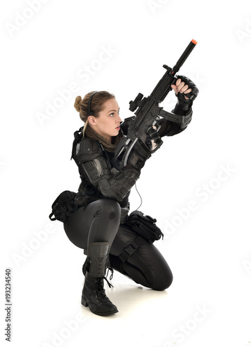 full length portrait of female wearing black tactical armour, crouching pose holding a weapon, isolated on white studio background.