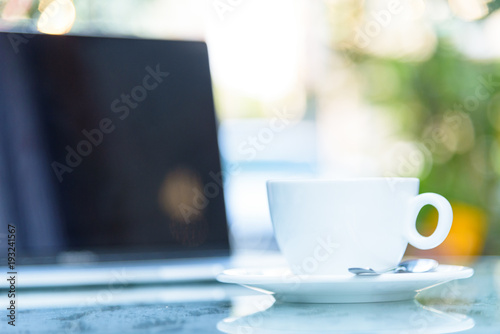A white cup of coffee and laptop ready to working in the morning with relaxing and yellow lighting bokeh background.