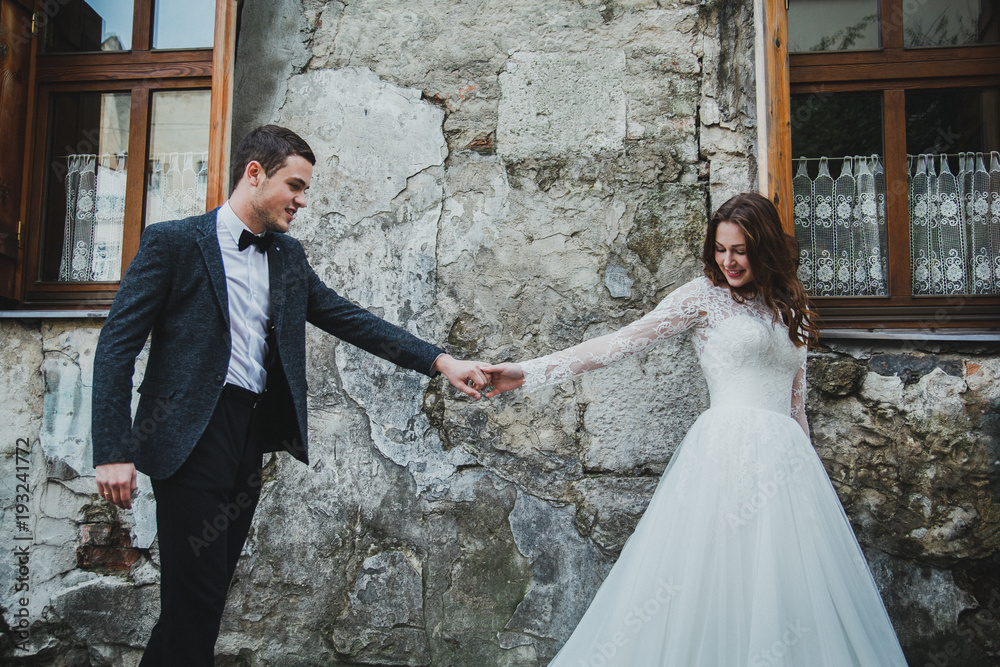 Wedding couple is walking in the old city. Stone walls in ancient sunny town on background. Rustic bride with hair down in lace dress and groom in grey suit and bow tie. Follow me photo. Wedding couple is walking in the old city. Stone walls in ancient sunny town on background. Rustic bride with hair down in lace dress and groom in grey suit and bow tie. Follow me photo.