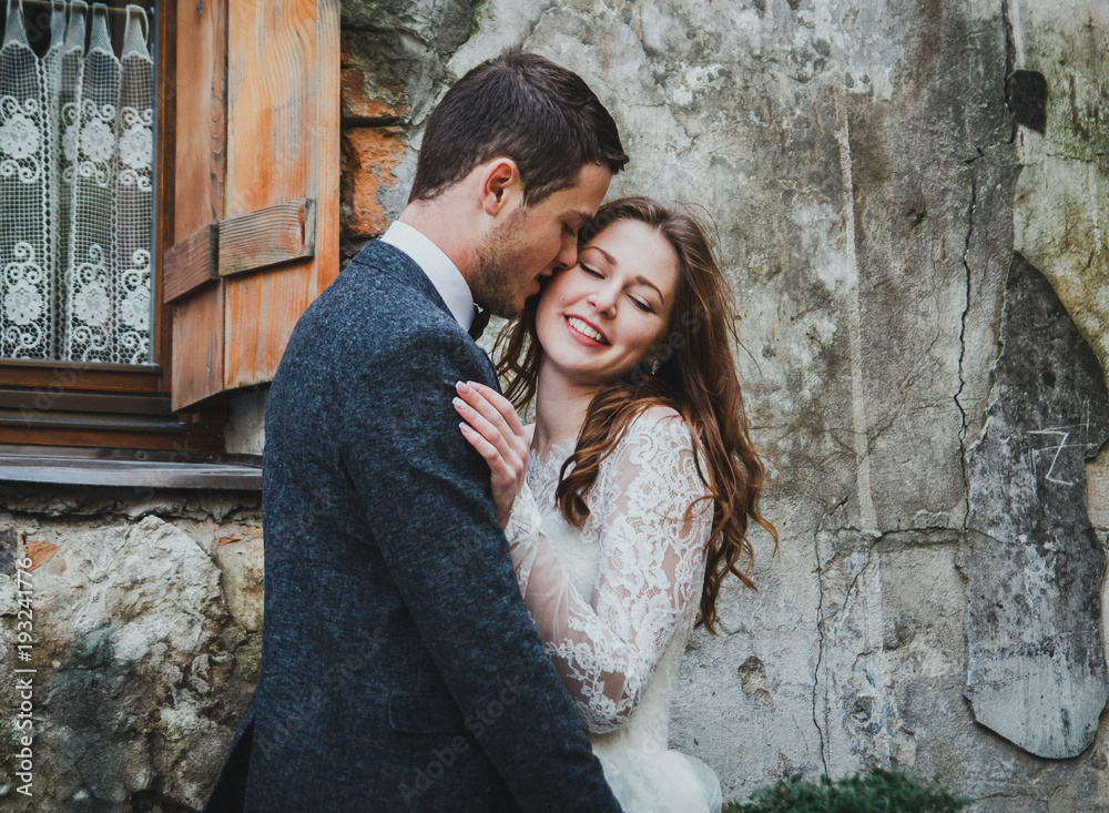 Wedding couple love story in old town. Groom is hugging the bride in white lace elegant dress. Hair down. Stone walls in ancient city. Fun and real emotions. Vintage atmosphere.