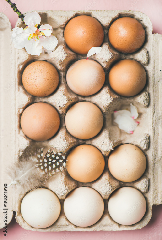 Easter holiday background. Flat-lay of gradient natural colored eggs in box with tender Spring almond blossom flower and feather over light pink background, top view. Greeting card concept