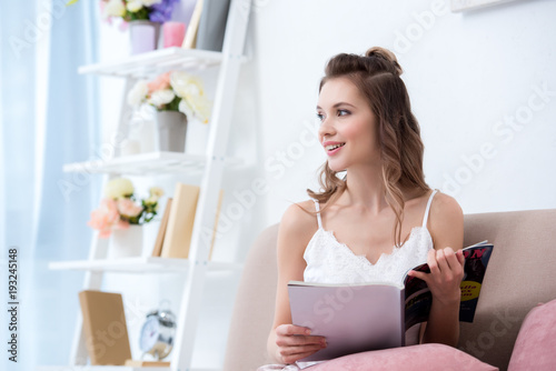 beautiful smiling girl holding fashion magazine and looking away at home
