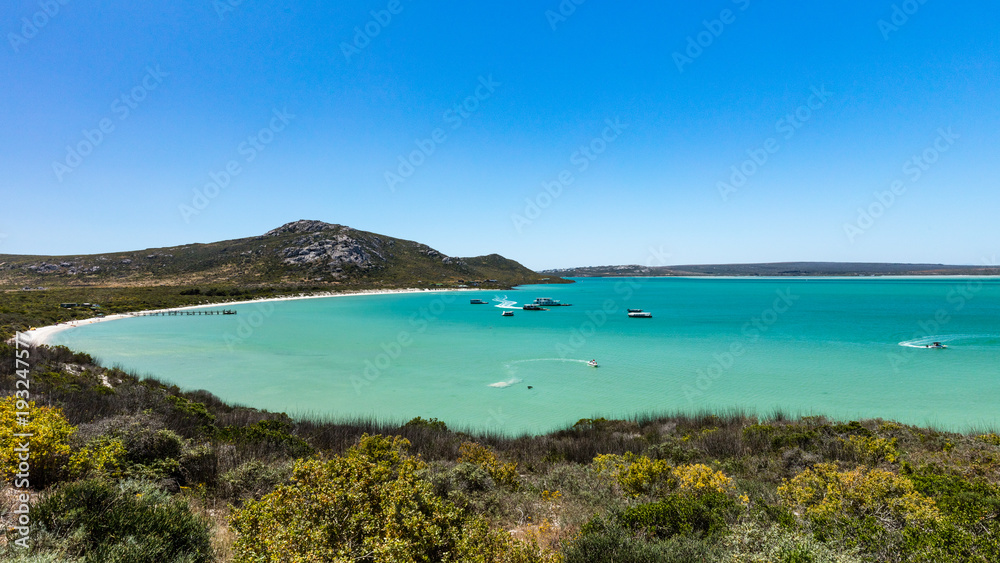 A view of Kraalbaai in the West Coast National Park in South Africa with boats leaving white trails in the sea. The sea in the bay is a beautiful turquoise colour and a clear blue sky